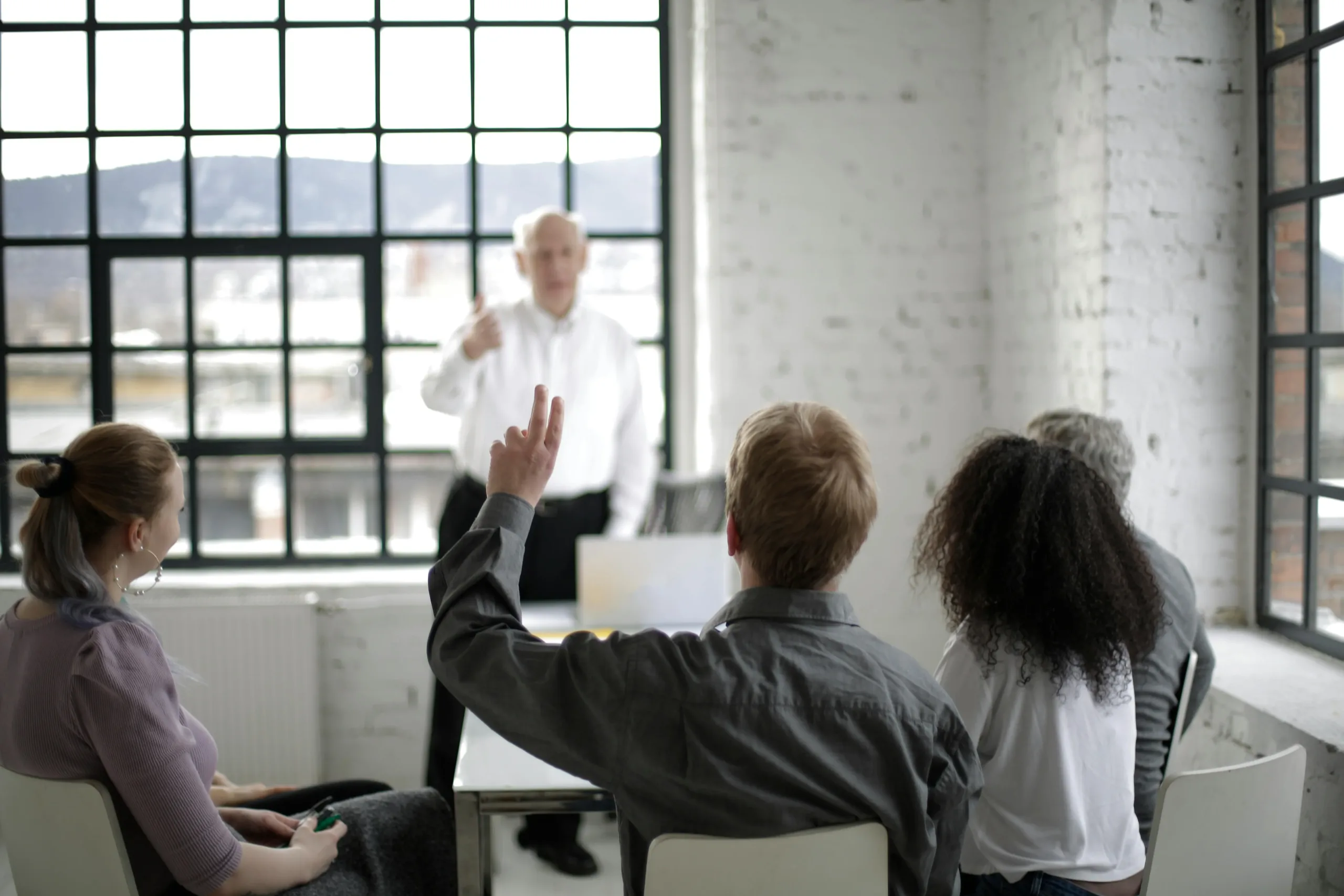 An accounting office or classroom. Three people are sitting, one person is standing at the front of the room. The person sitting in the middle has their hand raised, and the photographer is positioned behind them.