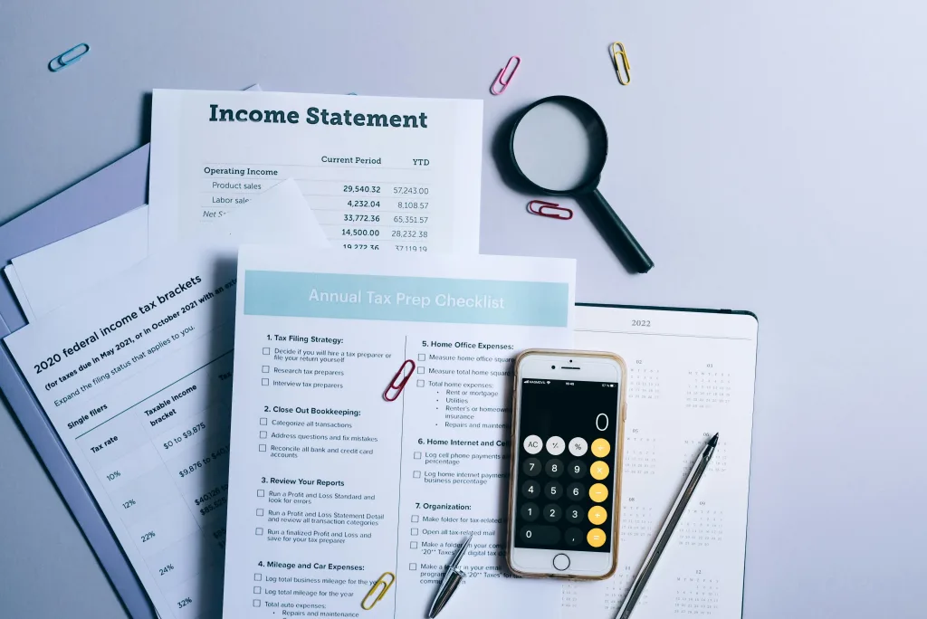 Various tax supplies are scattered across a desk, including paperwork, a calculator app open on an iPhone, magnifying glass, pencil, etc. 