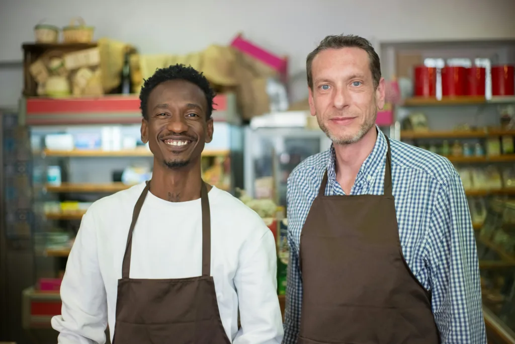 Two men wearing brown aprons, standing next to each other in a grocery store smiling at the camera. They will benefit from SALT deductions as small business partners.