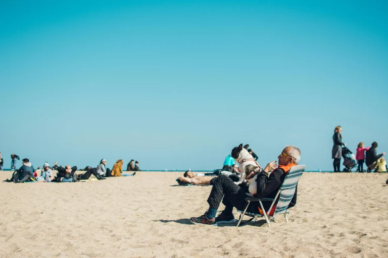 An elderly man is seated in a lounger with a small dog in his lap. They are at the beach in a sandy spot under a bright blue sky.