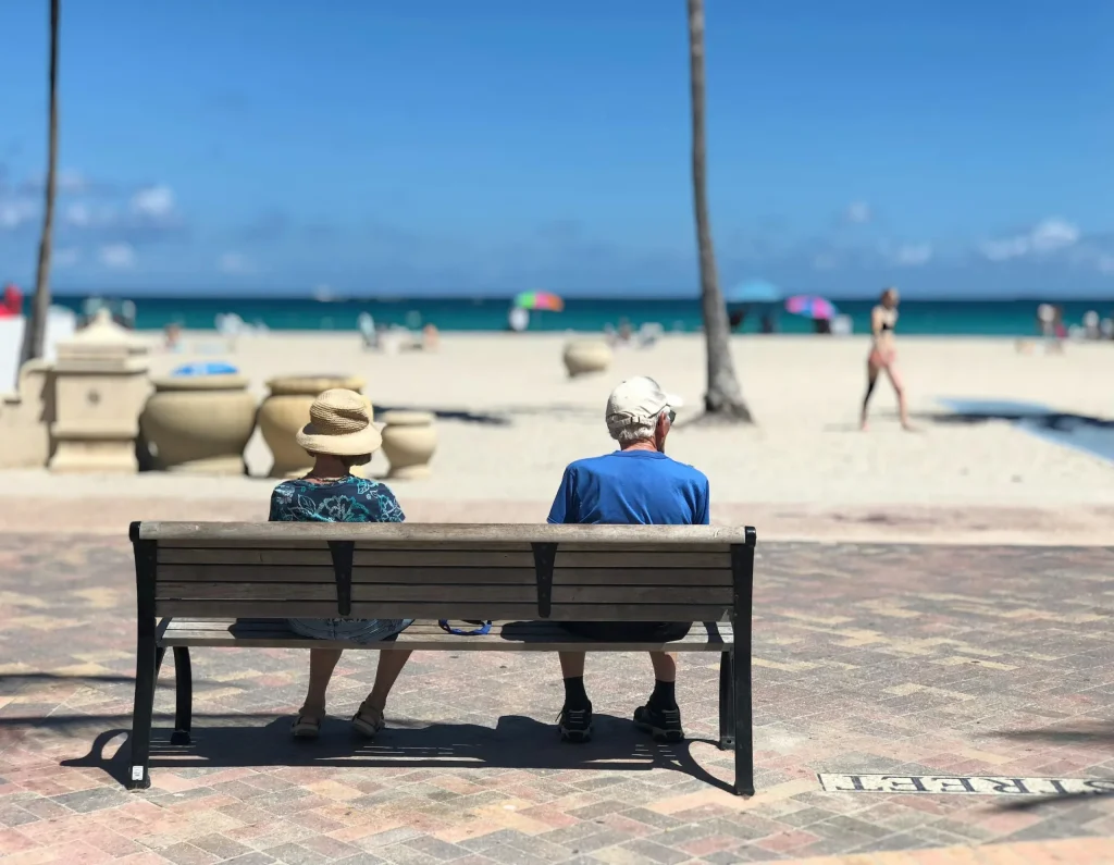 Pictured from behind, an elderly couple is sat down on a bench at the beach to discuss federal retirement under the big beautiful bill. 