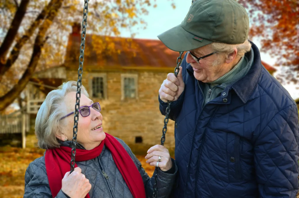 An elderly woman is on a swing, looking up at an elderly man who is holding the swing from the right side and smiling down at her.