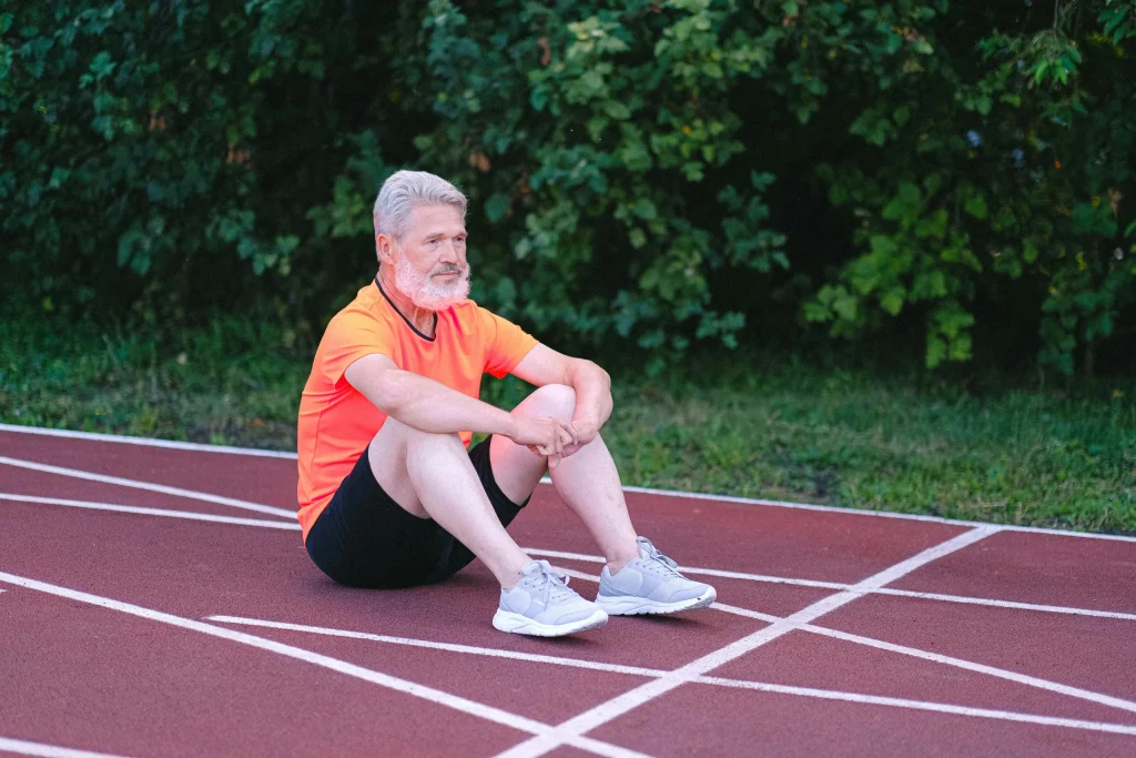 A grey-haired, bearded man dressed in running clothes is sitting on a race track.