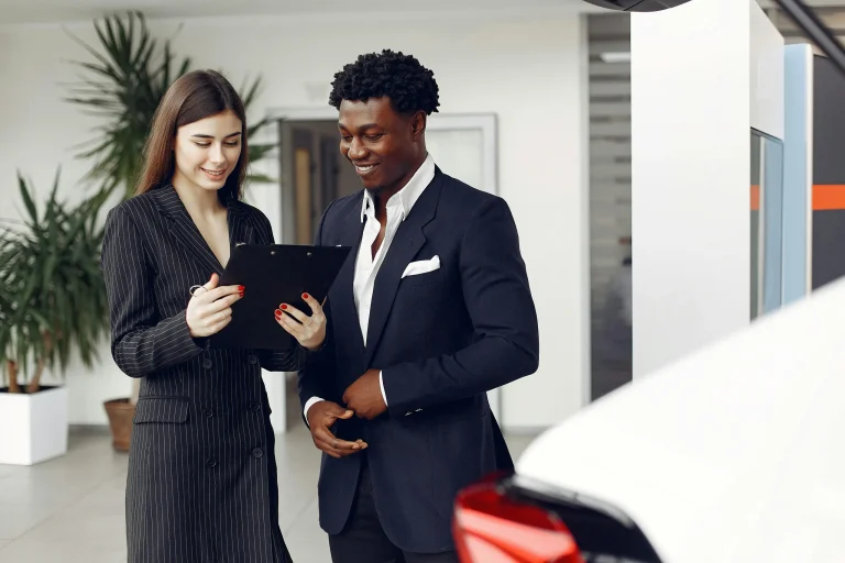 A business owner is leasing a car from a. dealership in California. They are standing indoors reviewing paperwork beside the car, which is in the foreground on the right side.