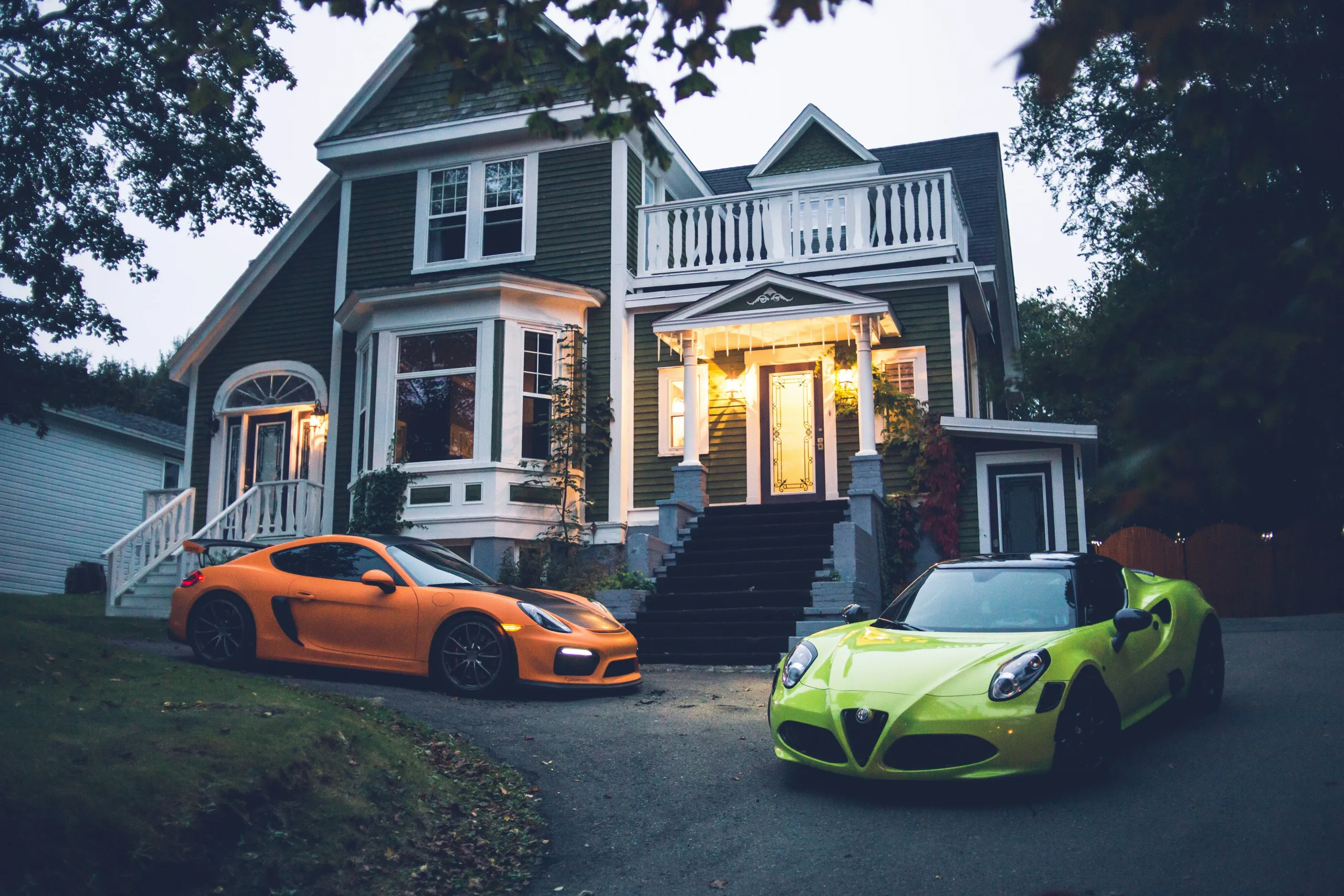 Two sports cars are parked in front of a nice Victorian style farmhouse.