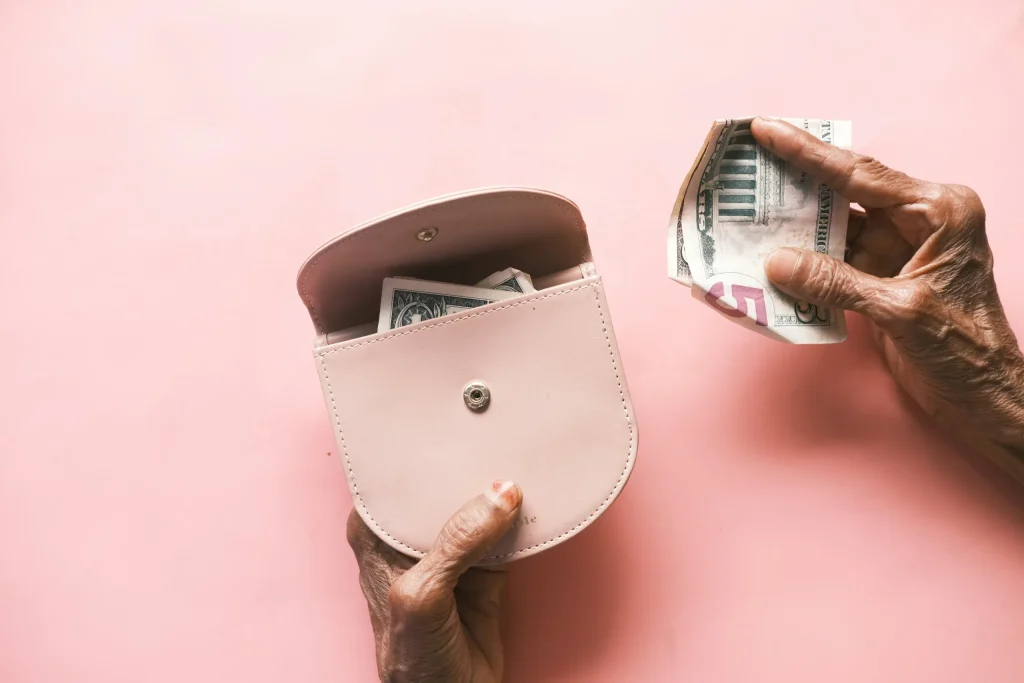 A photo of aged hands opening a white leather wallet and removing US cash. There is a pink background.